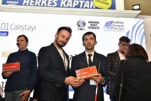 Fotoğraf, Ankara Kent Konseyi binasında bulunan konferans salonundaki sahnede çekilmiş. Arkada geniş bir dijital ekran bulunuyor ve ekranda "Ankara Kent Konseyi" ve "Kültür Sanat Meclisi" gibi logolar yer alıyor. Ayrıca "AKS101 Çalıştayı" ifadesi okunuyor. Üst kısımda, mavi bir pankartta "Herkes Kaptan" ifadesi yazıyor. Ön planda, resmi kıyafetler içinde bir grup insan yer alıyor. Sol tarafta, koyu mavi bir kazak giyen, boynunda bir mikrofon takılı olan ve plaket tutan Ali Caner Alpaslan var. Ortada, takım elbise giymiş ve kravat takmış bir kişi olarak ben varım. Elimde adımın yazdığı kırmızı bir plaket bulunuyor. Ayrıca diğer elimde beyaz baston var. Sağ tarafta açık renkli gömlek ve kareli bir ceket giymiş bir kişi duruyor. O kişi arka planda ve yüzü daha az belirgin. Fotoğraf karesine başka kişilerin girdiği de görülebiliyor.