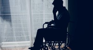 Bir tekerlekli sandalyede oturan adamın silueti. Adam, pencerenin önünde, perde arkasında oturuyor ve bir eliyle yüzüne dokunuyor. Oda karanlık, dışarıdan gelen ışık perdeyi ve adamın siluetini belirginleştiriyor. Adamın duruşu düşünceli veya üzgün bir hali yansıtıyor. Betimleme: Be My Eyes yapay zeka.