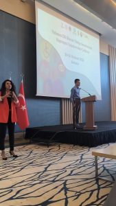 Bir konferans salonunda bir etkinlik sahnesi görülüyor. Sahnenin üzerinde büyük bir projeksiyon ekranı var. Ekranda "Yabancı Dil Olarak Türkçe Sınavlarında Kapsayıcı Uygulamalar Çalıştayı" yazıyor. Tarih olarak 23-25 Haziran 2025 ve yer olarak Ankara belirtilmiş. Sahnenin sağında, elimde beyaz baston bulunduğu halde kürsüde konuşuyorum. Sol tarafta ise kırmızı ceket giymiş bir kadın, işaret diliyle çeviri yapıyor. Arka planda Türkiye bayrağı yer alıyor. Salonun zemini açık renkli ve üzerinde koyu renk dallanmış desenler var. Betimleme: Be My Eyes yapay zeka.