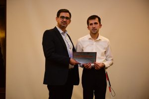 Sunum sonrası katılım belgesi alıyorum. Solda Oturum Başkanı koyu renk bir ceket, açık mavi gömlek ve gözlük takıyor. Sağda ise beyaz bir gömlek giymiş ve elimde beyaz baston tutar halde ben bulunuyorum. İkimiz birlikte, elimizde yatay olarak bir teşekkür belgesi tutuyoruz. Betimleme: Be My Eyes yapay zeka