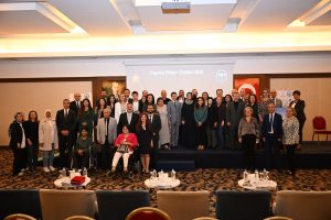 Engelsiz Bilişim 2025 Ödül Törenine ait toplu fotoğraf. Katılımcıların çoğu resmi giyimli; kadınlar ve erkekler birlikte, bazı katılımcılar tekerlekli sandalye kullanıyor ve çeşitli yaş gruplarından insanlar var. Sahnenin arka tarafında bir projeksiyon perdesinde "Engelsiz Bilişim Ödülleri 2025" yazısı gözüküyor. Yanlarda Türk bayrağı, Atatürk portresi ve çeşitli afişler bulunuyor. Ön planda ise yuvarlak masalarda su şişeleri, belgeler ve kırmızı bir kutu yer almakta, salonun zemininde desenli halı dikkati çekiyor. Katılımcılar kameraya dönük gülümseyerek poz vermişler. Betimleme: Be My Eyes yapay zeka