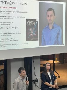 Toplantı salonunda düzenlenen panelde konuşma yaparken çekilen fotoğrafta, konuşmacılar olarak önde iki kişi sandalyede oturuyoruz. Solda ben elimde mikrofon tutar halde konuşuyorum. Sağda ise diğer panelist Özge Ünlü dinler vaziyette. Arkada, üzerinde “Emre Taşğın Kimdir?” başlıklı bir sunumun yansıtıldığı bir perde bulunuyor. Sunum slaytında (“Engellilik Posthümanizm ve Türkiye”) ve hakkımda çeşitli bilgiler yer almakta. Görselin arka planda masa ve sandalye detayları seçilebiliyor. Betimleme: Be My Eyes yapay zeka.