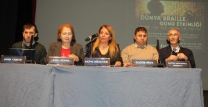 beş panelist, önümüzde isimlikler yer alıyor. Soldan sağa isimler: EMRE Taşgın, SİBEL GÜNEŞ, SAİME OĞUZHAN, ÖZGÜR UĞUR, FATİH MEHMET MORAY. Masanın üzerinde mavi bir örtü var. Ortada bir mikrofon bulunuyor. Arka planda ise bir afiş görünüyor; üstünde “DÜNYA BRAILLE GÜNÜ ETKİNLİĞİ” yazıyor ve Braille alfabesiyle 'Dünya Braille Günü Etkinliği' yazısı da var. Betimleme: Be My Eyes yapay zeka.

