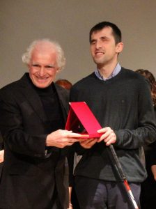 Plaket takdim anı. Sahnede ödülü veren kişi ile birlikte bir plaket tutuyoruz. Soldaki takım elbiseli kişi plaketi veriyor, sağda ise beyaz bastonum elimde olduğu halde kırmızı kutu içindeki plaketi alıyorum. Betimleme: Be My Eyes yapay zeka.