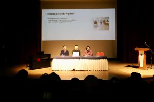 Kırmızı perdeli bir sahne üzerinde, arkasında 'Erişilebilirlik Nedir?' yazan bir projeksiyon ekranının önünde, uzun, beyaz ve lacivHert örtülü bir masada oturan üç panelistiz. Soldan sağa Ali Caner Alpaslan, ben ve Ayşegül yer alıyoruz. Önümde bilgisayarım var. Sahnenin solunda siyah bir piyano, sağında ise üzerinde mikrofon olan ahşap bir kürsü görülüyor. Betimleme: Gemini yapay zeka.