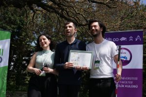 Güneşli ve ağaçlık bir alanda düzenlenen festivalde, sahnede Elimde teşekkür plaketi tutuyorum. Solumda Festival Koordinatörü Duru Arpacı elinde mikrofon tutuyor. Sağ tarafımızda ekipten Çağrı Sert arkadaşımız gülümsüyor. Beyaz bastonumu ona emanet ettiğim için elinde baston varken poz vermiş.