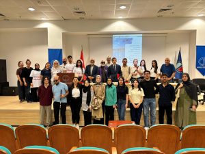 Anadolu Üniversitesi amfi sahnesinde, 'Engelliler Haftası' sunum perdesinin önünde poz veren kalabalık bir grubun toplu fotoğrafı. 