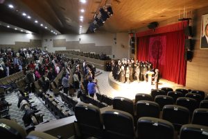 Etkinlikte konuşma yapan ve farklı görevleri bulunan kişilerin ayakta bulunduğu toplu fotoğraf. İzleyiciler oturur vaziyetteler.