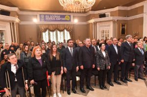 Ankara Kent Konseyi Engelliler Meclisi genel kurulunda, kalabalık bir grup salonun tavanından sarkan büyük kristal avize altında, sahne arkasındaki 'BU ŞEHİRDE HERKES KAPTAN' yazılı pankartın önünde duruyor. Betimleme: Gemini yapay zeka.