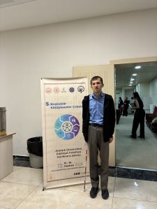 15-16 Haziran 2026 tarihlerinde Atatürk Üniversitesi Edebiyat Fakültesi Konferans Salonu'nda düzenlenen 5. Erişilebilir Kütüphaneler Çalıştayı'na ait ayaklı tanıtım afişinin yanında, elimde beyaz bastonumla ayakta duruyorum. Mavi gömlekli ve siyah ceketliyim.